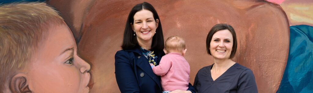 a woman and a baby and a nurse are smiling to camera in front of a mural of a woman breastfeeding a baby