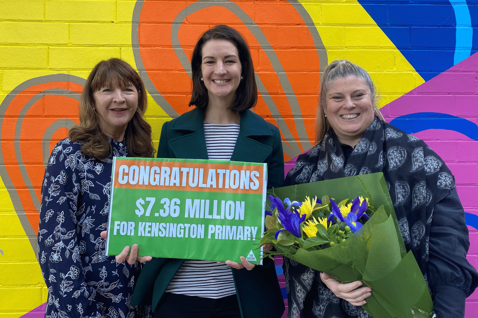 3 woman smiling to camera. One is holding a bunch of flowers and another is holding a sign that says: Congratulations $7.36 million for Kensington Primary